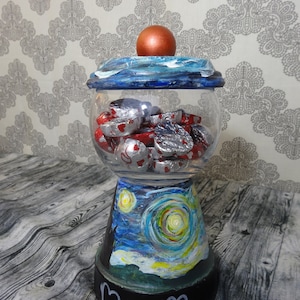 May include: A glass candy jar with a blue and white painted lid and base. The base is painted with a starry night scene. The jar is filled with silver and red foil-wrapped chocolates. The base of the jar has a black chalkboard label with the text "Mrs. M".
