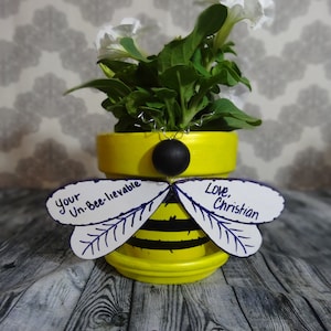 Puede incluir: Una maceta amarilla pintada para parecer una abeja con rayas negras y alas. Las alas tienen mensajes escritos a mano que dicen "Your Un-Bee-lievable" y "Love, Christian".