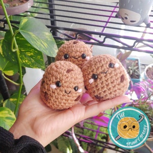 May include: Three crocheted brown cookie plush toys with cute faces. Each cookie has black eyes, a pink blush, and a smiling mouth. The cookies are sitting in a hand.