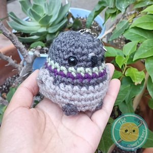 May include: A handmade crochet owl keychain in shades of gray, with black bead eyes and a decorative band of green and purple. The owl has a small silver keychain attached. The Grumpy Granny logo is visible in the background.