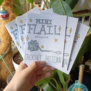 Könnte beinhalten: Eine Reihe weißer Mini-Flail-Schlüsselanhänger-Häkelmuster-Broschüren. Jede Broschüre zeigt den Titel in einer verspielten Schriftart, eine Cartoon-Flegel-Illustration und Sternakzente. Die Broschüren werden vor einer üppigen grünen Pflanze gehalten.