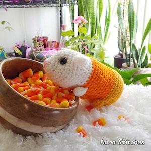 May include: A white and orange crocheted guinea pig wearing a candy corn costume. The guinea pig is sitting next to a bowl of candy corn.