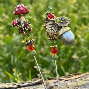 以下が含まれることがあります： 自然をモチーフにした2つの装飾ピン。1つは白い水玉模様の赤いキノコ、もう1つはドングリが特徴です。どちらのピンにも緑とオレンジの装飾ビーズが付いており、銀色のピンにセットされています。