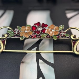 May include: A decorative metal bar with a floral design. The bar features three flowers in shades of red, orange, and yellow, with green leaves and small crystal accents. The bar has a gold finish and is likely used for decorative purposes.