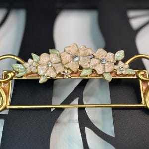 May include: A decorative gold-toned metal towel rack featuring a floral design. The rack has a horizontal bar for towels and is adorned with cream-colored flowers, green leaves, and sparkling crystal accents. The rack measures approximately 10 inches in width.
