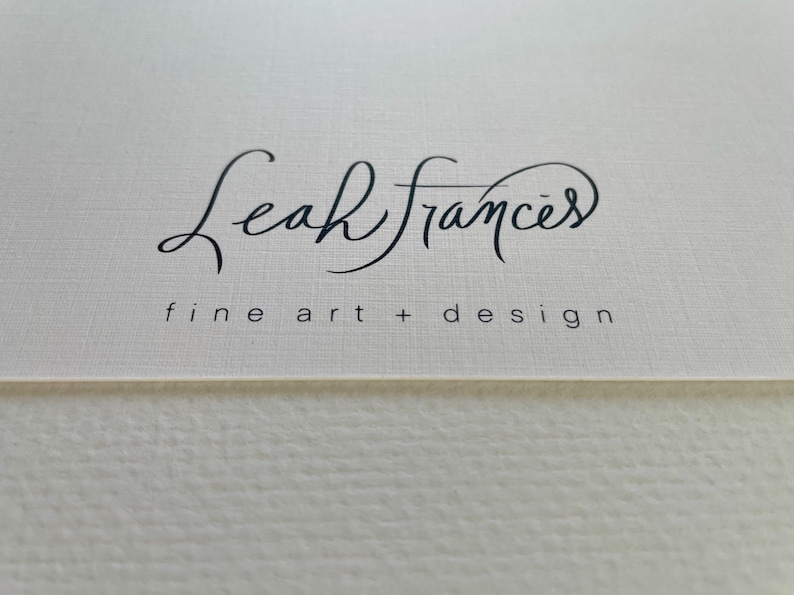 Puede incluir: Primer plano de una superficie texturizada blanquecina con el nombre "Leah Frances" en elegante escritura negra. Debajo del nombre, el texto dice "fine art + design" en una fuente m&aacute;s peque&ntilde;a, sin serifa.