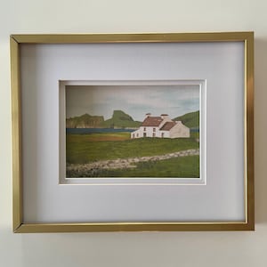 Puede incluir: Obra de arte paisaj&iacute;stica enmarcada que presenta una casa blanca con tejado rojo, situada frente a colinas verdes y un mar azul. La obra est&aacute; enmarcada en un marco met&aacute;lico dorado con un paspart&uacute; blanco.