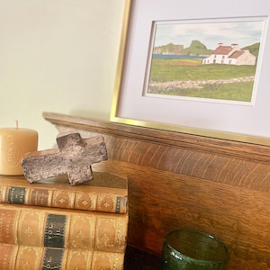 Puede incluir: Una pintura de paisaje enmarcada con un marco dorado, que representa una casa blanca con tejado rojo, campos verdes y un mar azul. Una pila de libros antiguos, una cruz de madera, un vaso de vidrio verde y una vela en primer plano.