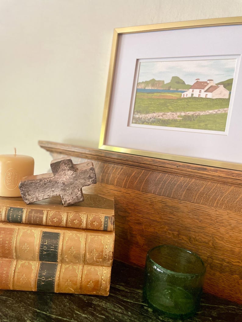 Puede incluir: Una pintura de paisaje enmarcada con un marco dorado, que representa una casa blanca con tejado rojo, campos verdes y un mar azul. Una pila de libros antiguos, una cruz de madera, un vaso de vidrio verde y una vela en primer plano.