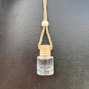 以下が含まれることがあります： 木製のキャップと吊り下げ用の織りロープが付いた、小さな透明なガラス瓶。ボトルは暗い背景に吊り下げられています。ロープには小さな木製のビーズと黄色のアクセントが付いています。