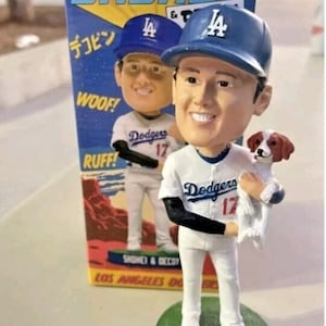 May include: A Shohei Ohtani bobblehead figure wearing a white Los Angeles Dodgers uniform with the number 17, holding a small brown and white dog. The figure stands on a green circular base labeled Shohei & Decoy, with its original box behind.