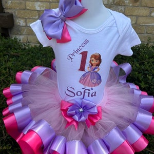 May include: A white onesie with the words "Princess 1 Sofia" and a picture of Princess Sofia from the Disney Junior show. The onesie is paired with a pink and purple tulle tutu with pink and purple satin ribbon.