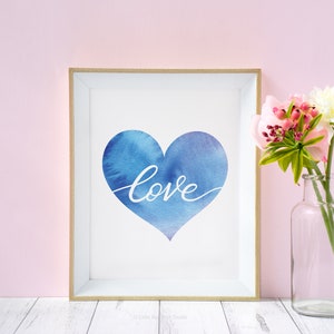 Könnte beinhalten: Ein gerahmter Druck mit einem blauen Aquarellherz und dem Wort "love" in weißer Kursivschrift. Der Druck wird auf einer weißen Oberfläche mit einer Vase mit rosa und weißen Blumen gezeigt.