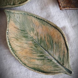 May include: A ceramic leaf-shaped dish with a textured surface. The dish features a detailed vein pattern and is painted in shades of green and tan. The Etsy shop name "FuzzyMudStudio.etsy.com" is visible at the bottom.