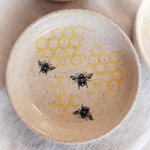 May include: A set of small, speckled ceramic dishes. Each dish features a yellow honeycomb pattern and black bee illustrations. The text "FuzzyMudStudio.etsy.com" is printed on the bottom of the image.