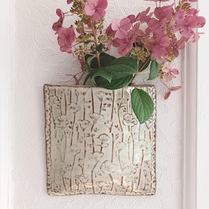 May include: A square ceramic wall pocket vase with a glossy off-white glaze and a brown etched floral pattern. The vase holds a bouquet of pink hydrangea flowers. The text FuzzyMudStudio.etsy.com is printed at the bottom of the image.