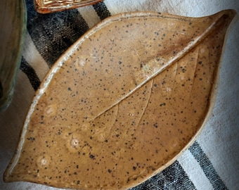 May include: A handmade ceramic leaf-shaped dish in a speckled brown glaze. The dish features a detailed vein pattern and is set on a striped fabric. The Etsy shop name "FuzzyMudStudio.etsy.com" is visible.