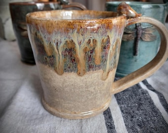 May include: A ceramic mug with a beige base and a glossy, dripping glaze in shades of brown, tan, and cream. The mug features a handle with a small sculpted dragonfly detail. The text FuzzyMudStudio.etsy.com is visible at the bottom.