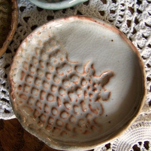 May include: A small, handmade ceramic dish with a light gray glaze and a textured, raised pattern resembling a cluster of grapes. The rim is a reddish-brown color. Other similar dishes are visible in the background.