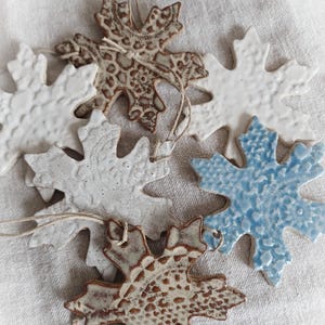 May include: A collection of ceramic leaf ornaments in various colors and patterns. Some are white, others are brown with a patterned design, and one is light blue. Each ornament has a string for hanging. The Etsy shop name "FuzzyMudStudio.etsy.com" is visible.