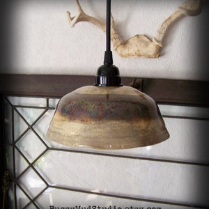May include: A brown and beige ceramic pendant light fixture with a black cord and socket. The light is hanging from a ceiling with a white textured wallpaper and a wooden beam.