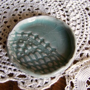May include: A small, round, ceramic dish with a light blue glaze. The dish features a textured, lace-like pattern on one side. The rim is slightly uneven. The dish is displayed on a white, crocheted doily.
