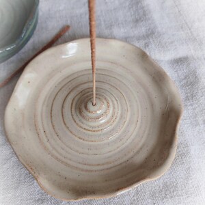 May include: A handmade, off-white ceramic incense holder with a spiral design. The holder has a shallow dish shape with a small hole in the center for the incense stick. The Etsy shop name "FuzzyMudStudio.etsy.com" is printed at the bottom.