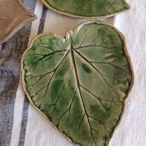 May include: Two ceramic dishes shaped like leaves with a glossy green glaze and visible vein textures. One dish is fully visible in the center, while another is partially shown at the top. The text FuzzyMudStudio.etsy.com is at the bottom.