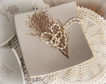 May include: A white ceramic dish with a brown floral design and a pearl necklace on top. The dish is sitting on a white crocheted doily.