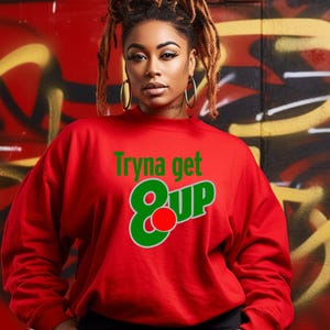 May include: Red sweatshirt with the text "Tryna get 8 Up" in green and red. The sweatshirt has long sleeves and a crew neck. The background is a red wall with graffiti.