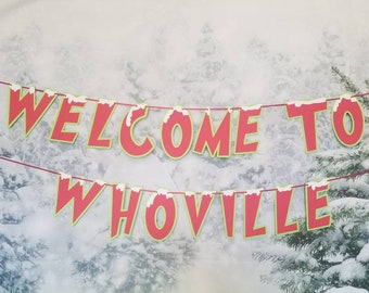 May include: A red and green banner with white snow accents reads "Welcome to Whoville". The banner is hanging in front of a snowy forest backdrop.