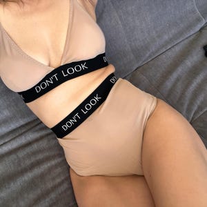 Peut inclure: Un ensemble de lingerie deux pièces beige composé d'une brassière triangle et d'une culotte taille haute. Les deux pièces présentent une large bande élastique noire avec le texte blanc DON'T LOOK imprimé sur le devant.