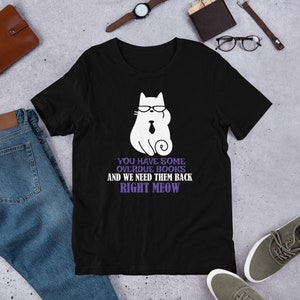 May include: Black t-shirt with a white cartoon cat wearing glasses and a tie. The text on the shirt reads "You have some overdue books and we need them back right meow."