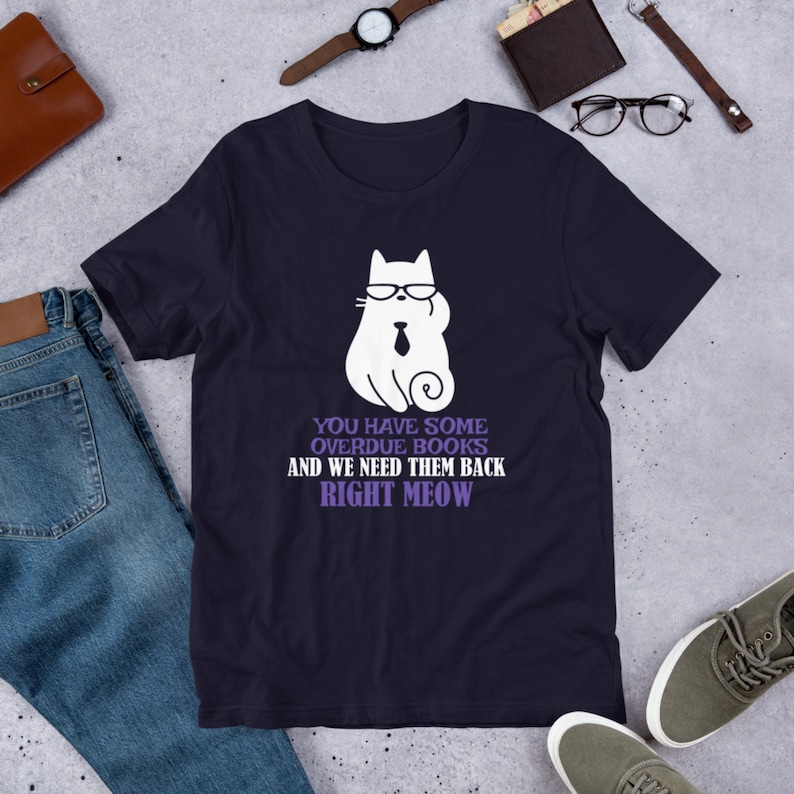 May include: A navy blue t-shirt with a white cat wearing glasses and a tie. The text on the shirt reads "You have some overdue books and we need them back right meow."