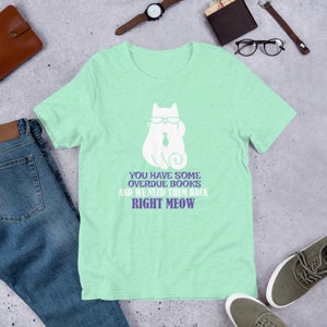 May include: A light green t-shirt with a white cat wearing glasses and a tie. The text on the shirt reads "You have some overdue books and we need them back right meow."