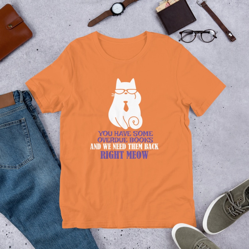 May include: Orange t-shirt with a white cartoon cat wearing glasses and a tie. The text on the shirt reads "You have some overdue books and we need them back right meow."