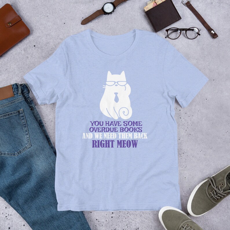 May include: Light blue t-shirt with a white cat wearing sunglasses and a tie. The text on the shirt reads "You have some overdue books and we need them back right meow."