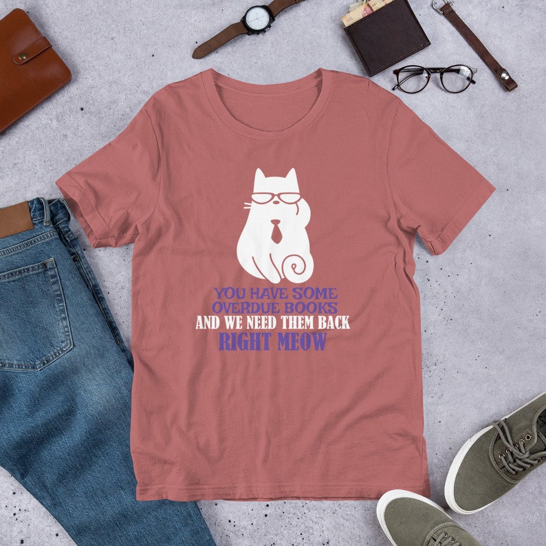 May include: A pink t-shirt with a white cat wearing sunglasses and a tie. The text on the shirt reads "You have some overdue books and we need them back right meow."