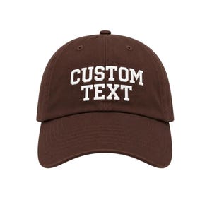 May include: A dark brown baseball cap featuring the words CUSTOM TEXT embroidered in white capital letters across the front. The cap has a structured crown, a curved brim, and a button at the top centre, providing a classic headwear design.