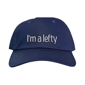 Pode incluir: Boné de beisebol azul marinho com aba curva. A frase "I'm a lefty" é bordada em branco na frente do boné. O boné tem um design clássico de seis painéis.