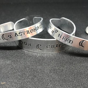 May include: Three silver bracelets with engraved text and crescent moon designs. The bracelets read "Future Astronaut", "Moon Child Need Space", and "Need Space".