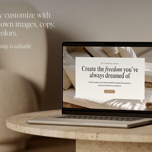 May include: A silver laptop shows a website mockup with the text "Create the freedom you've always dreamed of." The laptop is on a round, light-coloured stone table. The background is a neutral beige.