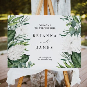 May include: A white wedding welcome sign with green foliage and white flowers. The sign reads "Welcome to our wedding Brianna and James The Second of August Two Thousand and Twenty".