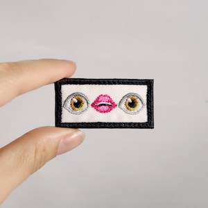 May include: White embroidered patch with a pair of eyes and a pink mouth. The patch is surrounded by a black border.