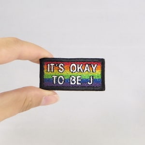 May include: A rainbow-colored embroidered patch with the text "It's okay to be J" in white lettering.