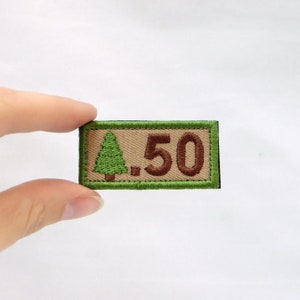 May include: A rectangular patch with a green border and a tan center. The patch features a green embroidered tree on the left and the number "50" in brown on the right. The patch is being held by a person.