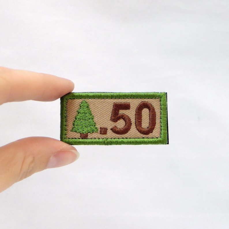 May include: A rectangular patch with a green border and a tan centre. The patch features a green embroidered tree on the left and the number "50" in brown on the right. The patch is being held by a person.
