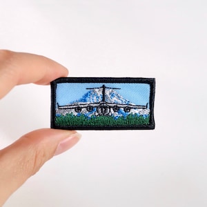 May include: Embroidered patch featuring a detailed image of an airplane flying over a mountain range. The patch has a blue sky background, green grass, and a black border. The airplane is silver and the mountain is white.