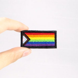 Könnte beinhalten: Gestickter rechteckiger Aufnäher mit dem Design der Progress Pride Flagge. Die Flagge hat einen schwarzen Rand und enthält die traditionellen Regenbogenstreifen sowie einen Chevron aus Farben, die Transgender, Marginalisierte und People of Color repräsentieren.