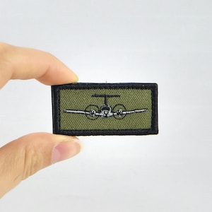 May include: A rectangular patch with a green background and a black border. The patch features a detailed embroidered image of a twin-engine airplane in white and black. The patch is held by a person's fingers.